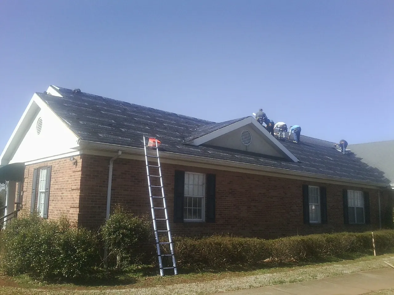 Crew tearing shingles off a brick ranch roof with a ladder set — Metal Roof Repair work by our crew in Rosenberg