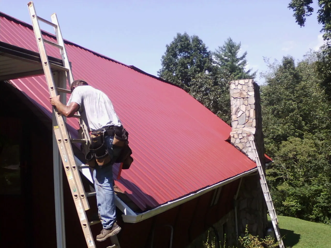 Roofer on a ladder beside a red metal roof and stone chimney — Soffit & Fascia Repair by Roofing Brothers in Rosenberg